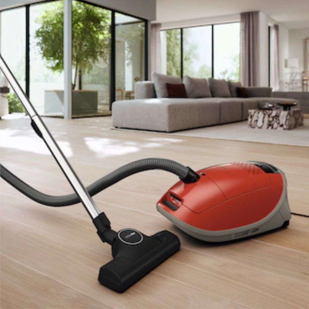 Miele cylinder vacuum cleaner on wooden flooring in a bright living room.