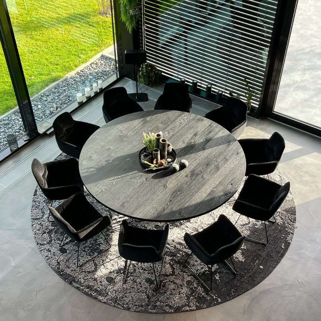 miinu round patterned rug framing an eight-seat black dining set in a glass-walled room.