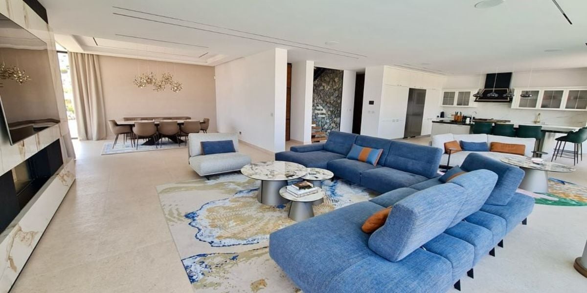 Large luxury open plan living room with blue sectional sofas, statement rug, marble coffee tables, dining area and open kitchen beyond