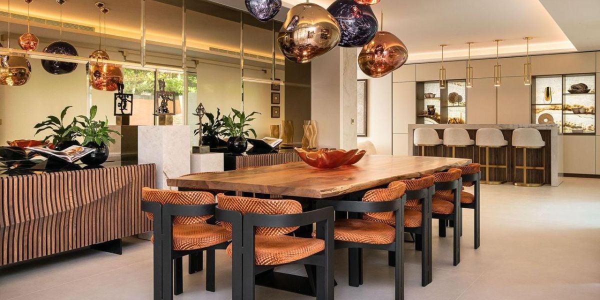 Bold open plan dining kitchen with live-edge wood table, orange upholstered chairs, copper and bronze pendant cluster and integrated bar seating beyond