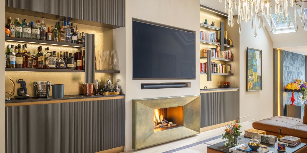 Open plan living room with integrated home bar built into dark ribbed joinery, brass-edged fireplace, open shelving and crystal chandelier above