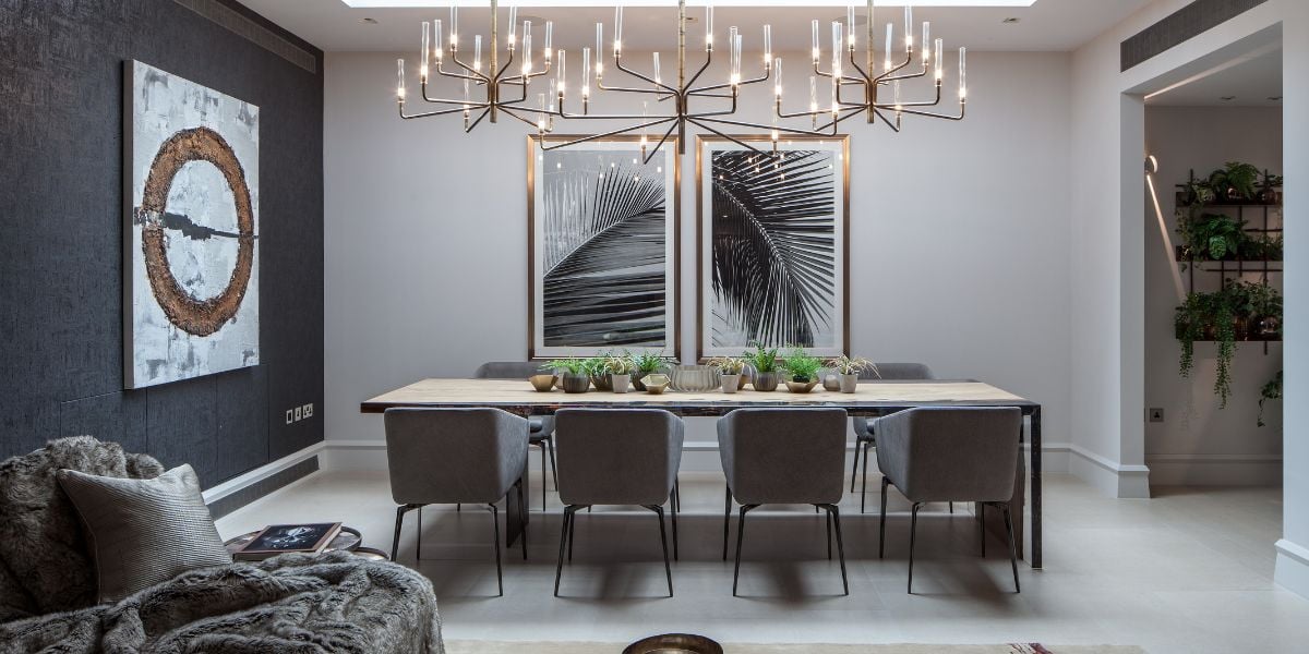 Elegant open plan dining room with dramatic multi-arm brass chandelier, long dining table with grey velvet chairs, botanical large-format artwork and dark textured feature wall