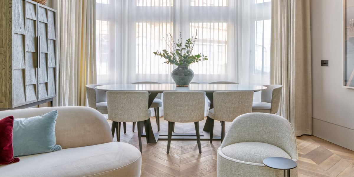 Small open plan living and dining room with herringbone oak floor, oval dining table, rounded upholstered armchairs and sheer floor-to-ceiling curtains